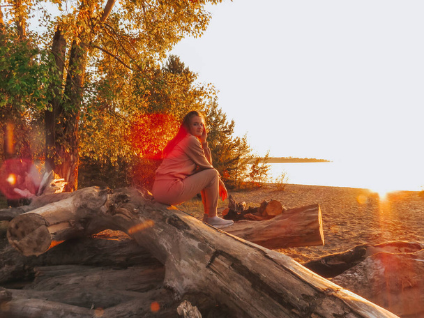 red-haired young woman in a beige sports suit in the light of orange sun glare at sunset on the seashore on a pile of dry tree trunks against the background of yellow autumn leaves, emotions of joy, calm, happiness - Fotografie, Obrázek