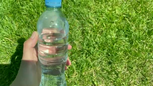 El, parlak güneş ışığında yeşil çimenlerin arka planında bir şişe taze su tutuyor. Açık hava sporu yapıyorum. Sıcak hava konseptinde susuzluk ve su dengesi. Plastik şişede su. - Video, Çekim