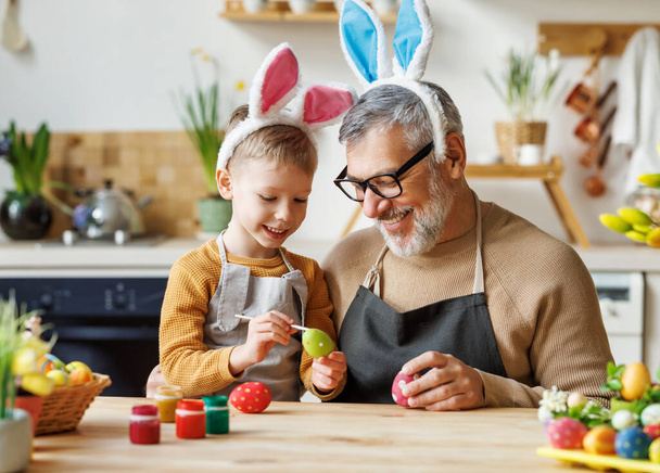 Liefdevolle grootvader en kind in konijnenoor hoofdbanden versieren eieren voor Pasen in lichte gezellige keuken - Foto, afbeelding