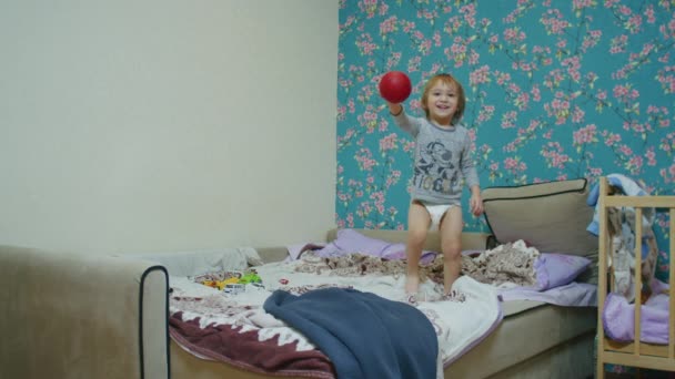 un pequeño niño feliz salta en la cama en casa y juega pelota con su padre, el padre le lanza la pelota fuera de la pantalla. - Metraje, vídeo