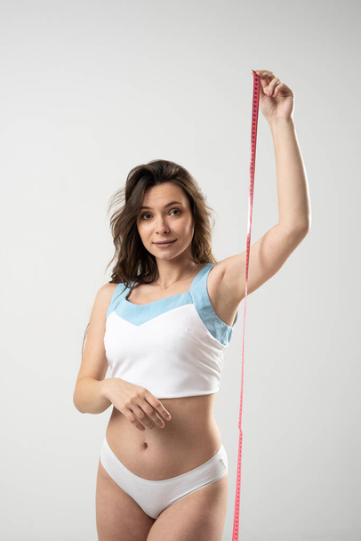 Confused brunette woman with slim body in white underwear on a diet looking in a camera and showing a pink centimeter tape. Healthy lifestyles concept. Sport and diet. - Zdjęcie, obraz