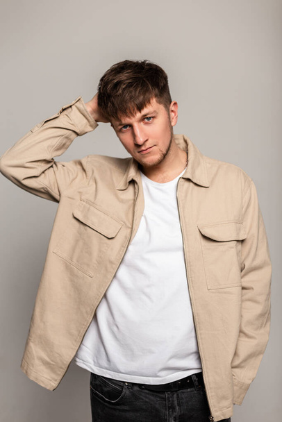Мода модель молодой человек с прической в модной наружности с бежевой курткой и белой футболкой позирует в студии на сером фоне - Фото, изображение