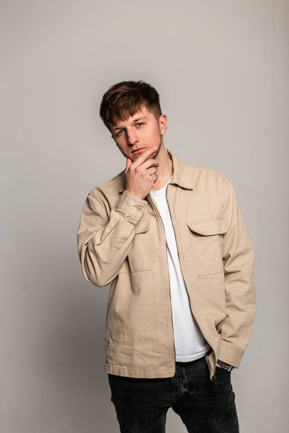 Havalı bej ceketli ve beyaz tişörtlü şık, yakışıklı genç adam stüdyoda gri arka planda poz veriyor. Erkek güzelliği ve modası - Fotoğraf, Görsel