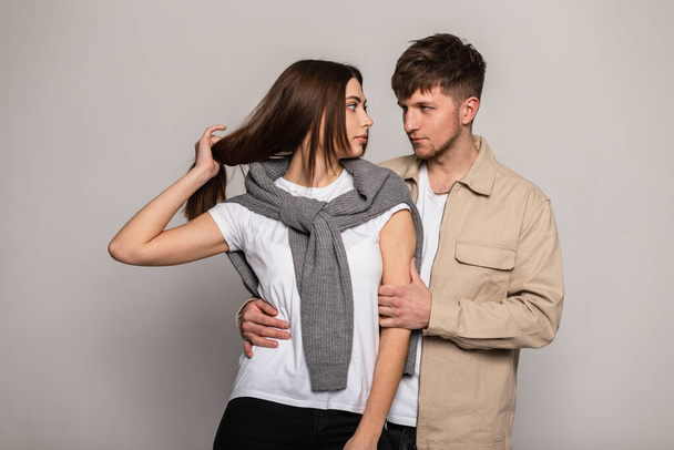 Handsome young fashionable couple woman and handsome man in stylish outerwear with white t-shirts, vintage sweater and beige jacket in studio on gray background - Fotó, kép