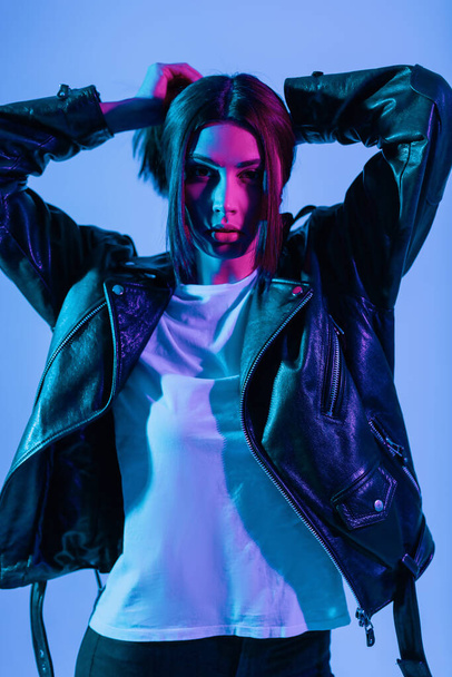 Pretty young stylish girl model in a fashionable black retro leather jacket with a white T-shirt straightens her hair in the studio with creative neon pink light. Colored portrait of a girl - Φωτογραφία, εικόνα
