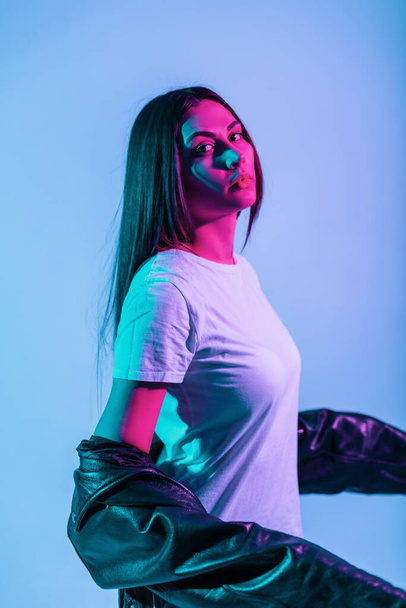 Fashionable pretty girl with a cute face in fashionable clothes with a leather black jacket and a white T-shirt posing in the studio with pink and blue lights. Creative portrait of a beautiful woman - Фото, зображення
