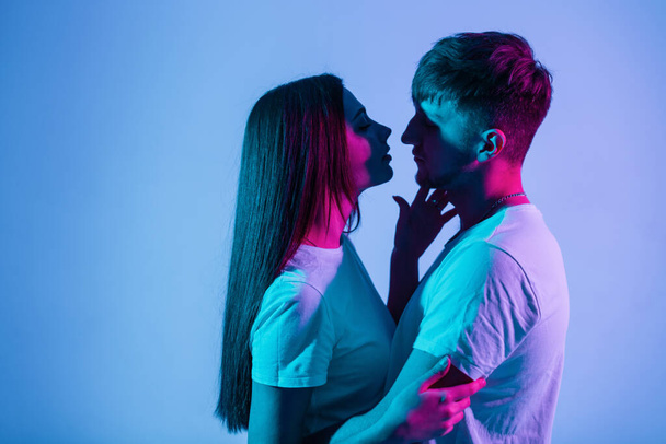 Beautiful couple of people in love with creative colorful blue and pink light. Pretty fashionable woman with handsome stylish man in white t-shirts kissing in studio - Fotoğraf, Görsel