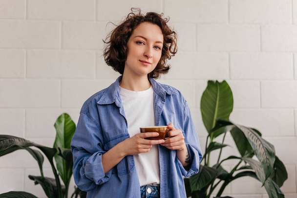 Smiling young woman with short curly hair at cafe drinking coffee in front of green leaves indoors - 写真・画像