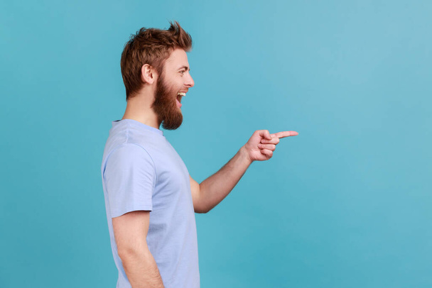 Side view of positive satisfied bearded man pointing aside, showing blank copy space for idea presentation, commercial text, smiling happily. Indoor studio shot isolated on blue background. - Φωτογραφία, εικόνα
