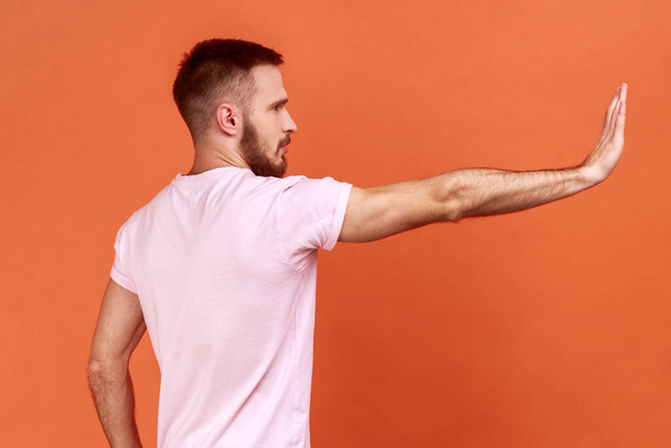 Side view of serious man making stop gesture showing palm of hand, conflict prohibition warning about danger, stop bullying, wearing pink T-shirt. Indoor studio shot isolated on orange background. - Foto, Imagen