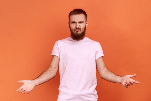 Portrait of angry bearded man standing with raised hands and surprised indignant expression, asking why how, what reason, wearing pink T-shirt. Indoor studio shot isolated on orange background. - Photo, Image