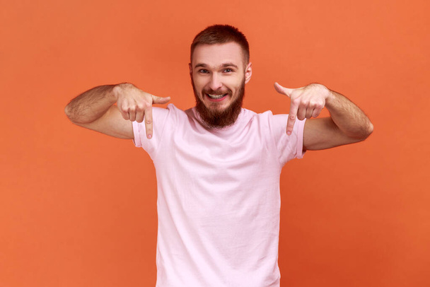 Cheerful satisfied man pointing fingers down, paying attention to advertising area, blogger recommending to subscribe, wearing pink T-shirt. Indoor studio shot isolated on orange background. - Photo, Image