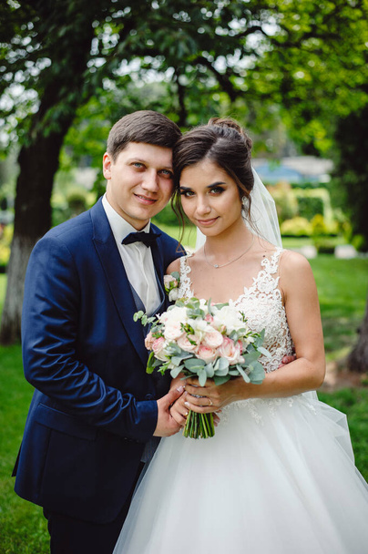 Portrait of romantic couple of newlyweds, groom in blue tuxedo and bride in white dress stands and holds a beautiful bouquet of pink and white peony flowers. Wedding ceremony in nature. - Фото, изображение