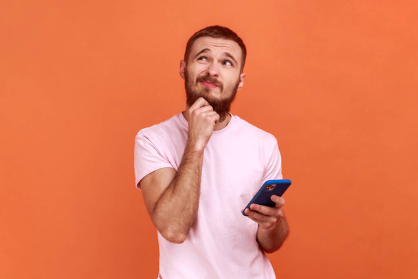 Portrait of man holding chin, standing with smartphone in hand, contemplating about software updating, choosing suitable tariffs, wearing pink T-shirt. Indoor studio shot isolated on orange background - Foto, Bild
