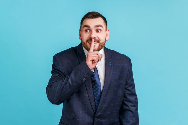 Portrait of businessman wearing official style suit holding finger near lips showing shh gesture, keeping secrets preparing surprise. Indoor studio shot isolated on blue background. - Foto, afbeelding