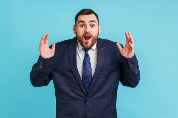 Wow, it's unbelievable! Surprised bearded man in official style suit staring at camera with widely open mouth and big eyes, raising arms in amazement. Indoor studio shot isolated on blue background. - Фото, изображение