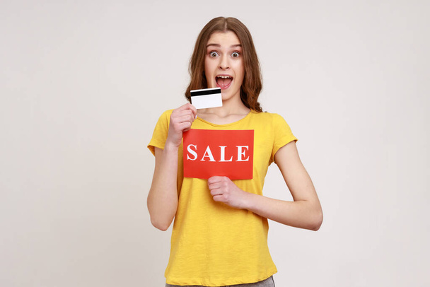 Cheerful excited woman in yellow T-shirt holding showing credit card and card with sale inscription, pay for purchases with credit card, banking. Indoor studio shot isolated on gray background. - Фото, зображення