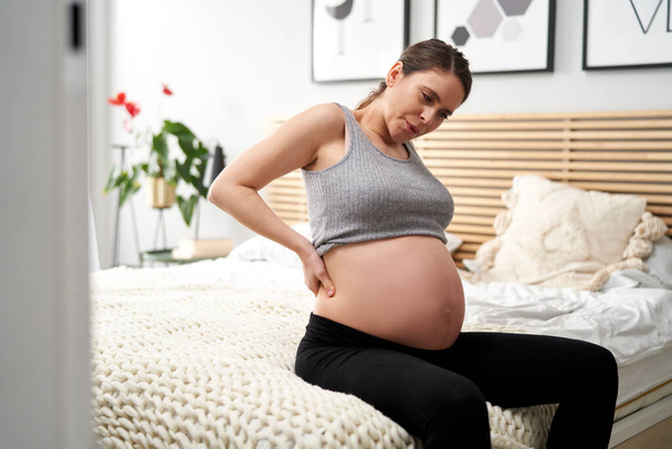 Kaukasierin in fortgeschrittener Schwangerschaft sitzt auf Bett und hat starke Krämpfe und Rückenschmerzen  - Foto, Bild