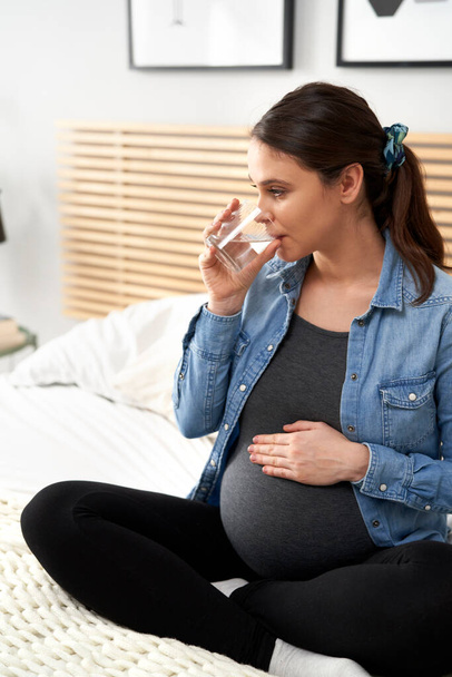 Pystysuora kuva valkoihoinen nainen pitkälle raskauden istuu sängyllä ja juomaveden - Valokuva, kuva