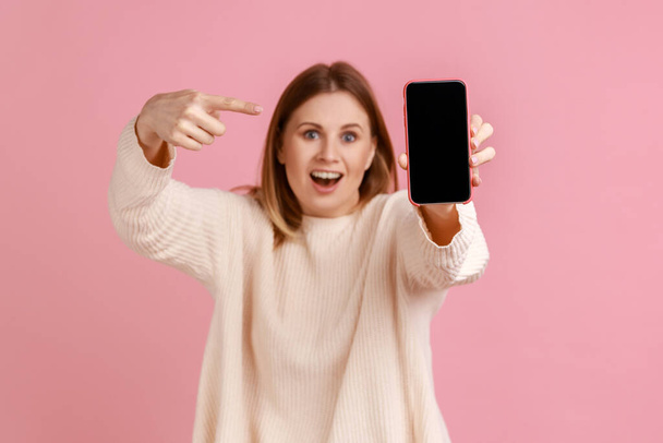 Portrait of shocked woman holding out smartphone, pointing at device blank display with copy space for promotional text or ad, wearing white sweater. Indoor studio shot isolated on pink background. - Foto, afbeelding