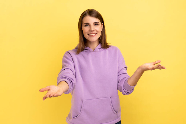 Openhearted generous woman outstretching hands looking at camera with kind smile, greeting and regaling, happy glad to see you, wearing purple hoodie. Indoor studio shot isolated on yellow background. - Foto, immagini