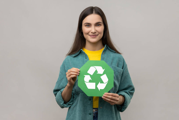 Smiling woman with dark hair holding in hands green recycling sing, ecology concept, looking at camera, wearing casual style jacket. Indoor studio shot isolated on gray background. - Foto, Imagen