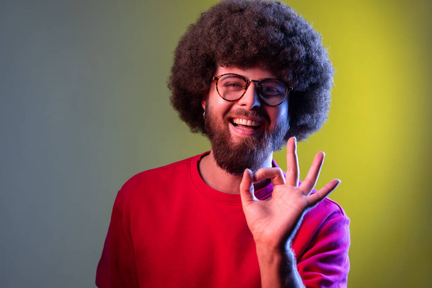 Happy smiling hipster man with Afro hairstyle showing okay sign, looking at camera with positive expression, wearing red sweatshirt. Indoor studio shot isolated on colorful neon light background. - Fotografie, Obrázek