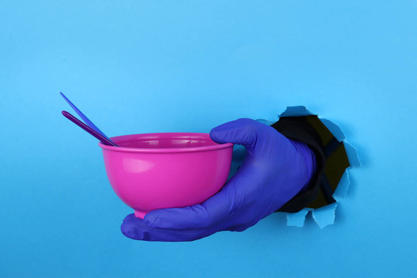 hand in glove holds hair dye bowl. A hole in the blue paper. Hair coloring in the salon or at home - Valokuva, kuva