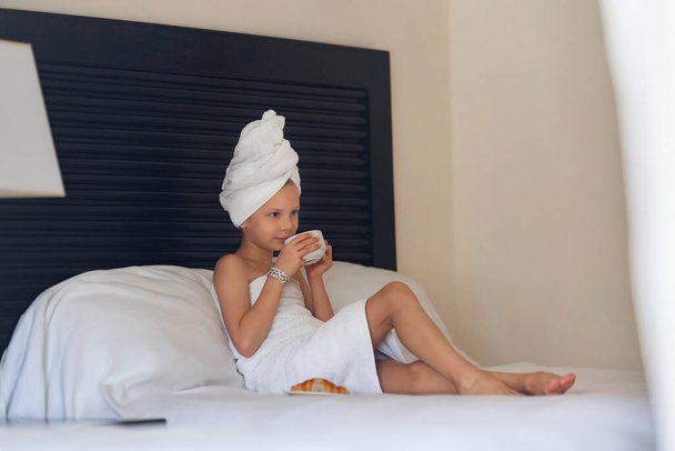 A little girl in a white towel drinks morning coffee in bed at home - Foto, Imagen