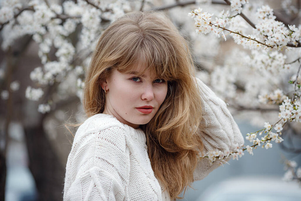 красивая молодая женщина в белом свитере в саду с сакурой, весеннее время. открытая мода - Фото, изображение
