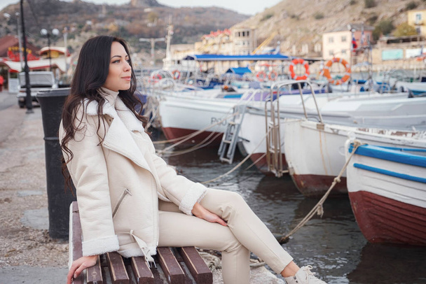 Eine unbeschwerte kaukasische Frau in beiger Kleidung genießt den Blick auf das Meer an einem warmen, windigen Tag. Porträt vor einer Meeresbucht mit Booten auf der Seebrücke - Foto, Bild