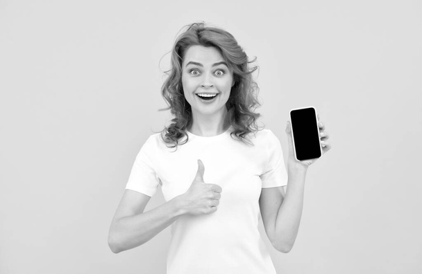 opgewonden jonge vrouw geïsoleerde overgele achtergrond met behulp van mobiele telefoon tonen ok gebaar. - Foto, afbeelding