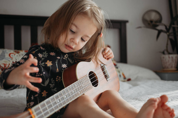 toddler girl playing ukulele guitar sitting on the bed at home - Foto, afbeelding