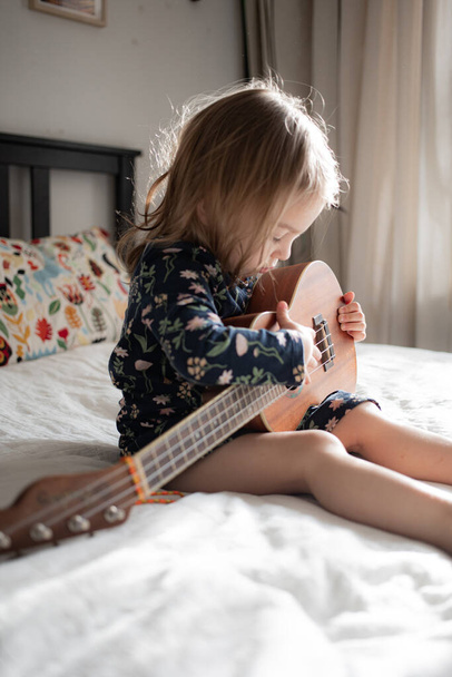 peuter meisje spelen ukelele gitaar zitten op het bed thuis - Foto, afbeelding