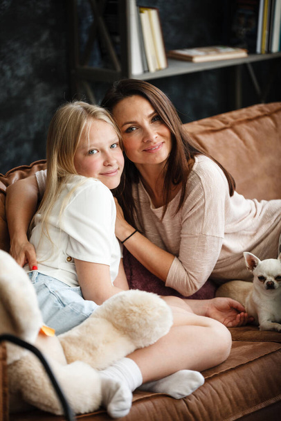 Mother and daughter relationship concept. A woman and a teenage girl are sitting in front of the TV on the couch and have fun talking. The family spends time at home. Little dog lying on laps - Photo, Image