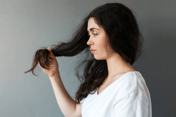 Portrait of young caucasian sad woman looks at the tips of her long dark hair. Gray background. Side view. The concept of hair care. - Photo, Image