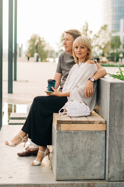 An adult mature happy couple in love sitting on bench outdoors in city street park. A blonde caucasian man and woman spend time together and drinking coffee. Senior wife and husband walking outside. - Фото, зображення
