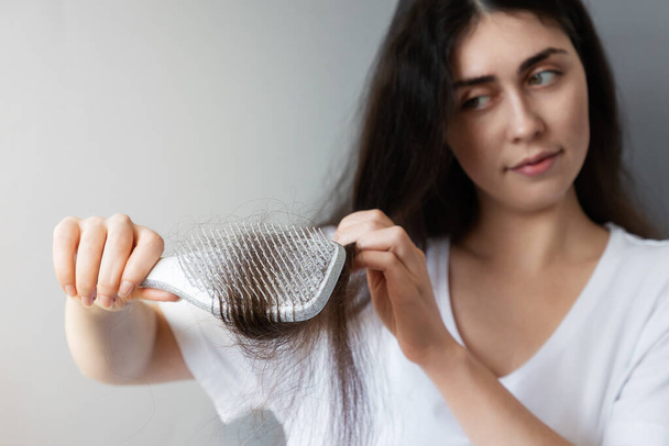 Retrato de uma jovem caucasiana penteando seu cabelo escuro emaranhado. Close up de escova de cabelo com perda de cabelo. Fundo azul. O conceito de calvície. - Foto, Imagem