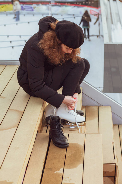Una joven se pone patines de hielo en los pies, sentada en escalones de madera. - Foto, Imagen