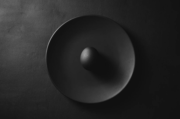 Top view of one black egg in a plate on the dark moody background. Minimal easter concept. - Zdjęcie, obraz