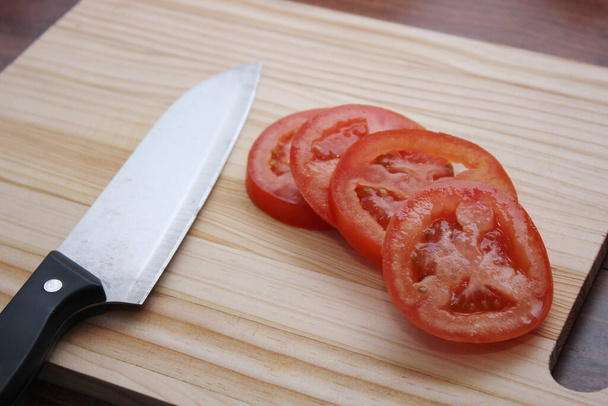 Tomates fatiados e facas são colocados em uma tábua de corte de madeira em uma mesa de madeira. - Foto, Imagem