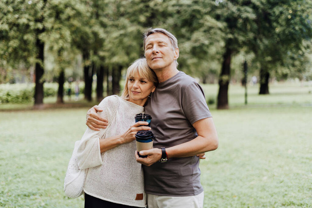 Ein erwachsenes, glückliches verliebtes Paar, das sich im Stadtpark umarmt. Ein blonder kaukasischer Mann und eine Frau verbringen Zeit miteinander und trinken Kaffee. Senior Frau und Mann zu Fuß nach draußen. - Foto, Bild