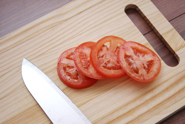 Pomidory w plasterkach i noże umieszczone są na drewnianej desce do krojenia w drewnianym stole.. - Zdjęcie, obraz