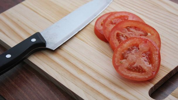 Pomidory w plasterkach i noże umieszczone są na drewnianej desce do krojenia w drewnianym stole.. - Zdjęcie, obraz