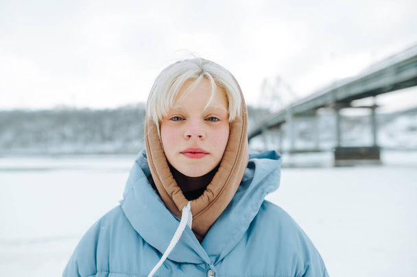 schönes Mädchen mit weißen Augenbrauen und Haaren in blauer Jacke steht im Winter draußen in der Kälte und blickt in die Kamera, Nahaufnahme Porträt - Foto, Bild