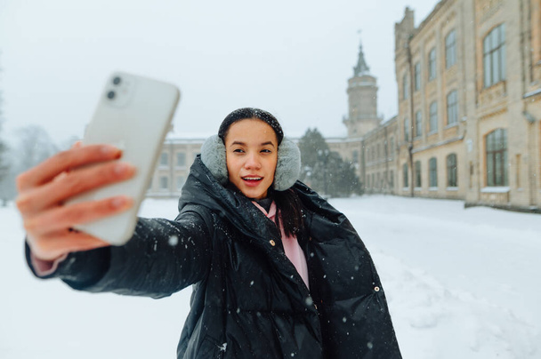 Dáma si bere zimní selfie ve sněžném dni. Krásná žena v teplém kabátě a kapuci stojí na ulici ve sněhu a vezme selfie na kameru s chytrým telefonem. - Fotografie, Obrázek
