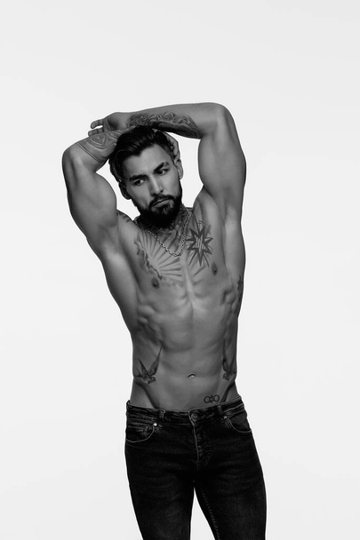 Latino shirtloos mannelijk model met gespierde getatoeëerde romp staande met handen in zakken en wegkijkend op grijze achtergrond - Foto, afbeelding