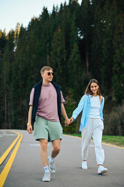 Élégant jeune couple homme et femme marchant main dans la main sur une route forestière lors d'un voyage et souriant. Vertical. - Photo, image