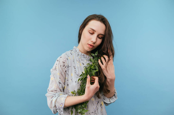 Atrakcyjna kobieta w sukience z kręconą rośliną w dłoniach odizolowana na niebieskim tle, delikatnie przytulająca roślinę. - Zdjęcie, obraz