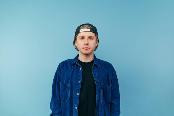 Handsome young man in denim shirt and cap isolated on blue background, looking at camera with serious face. - Zdjęcie, obraz